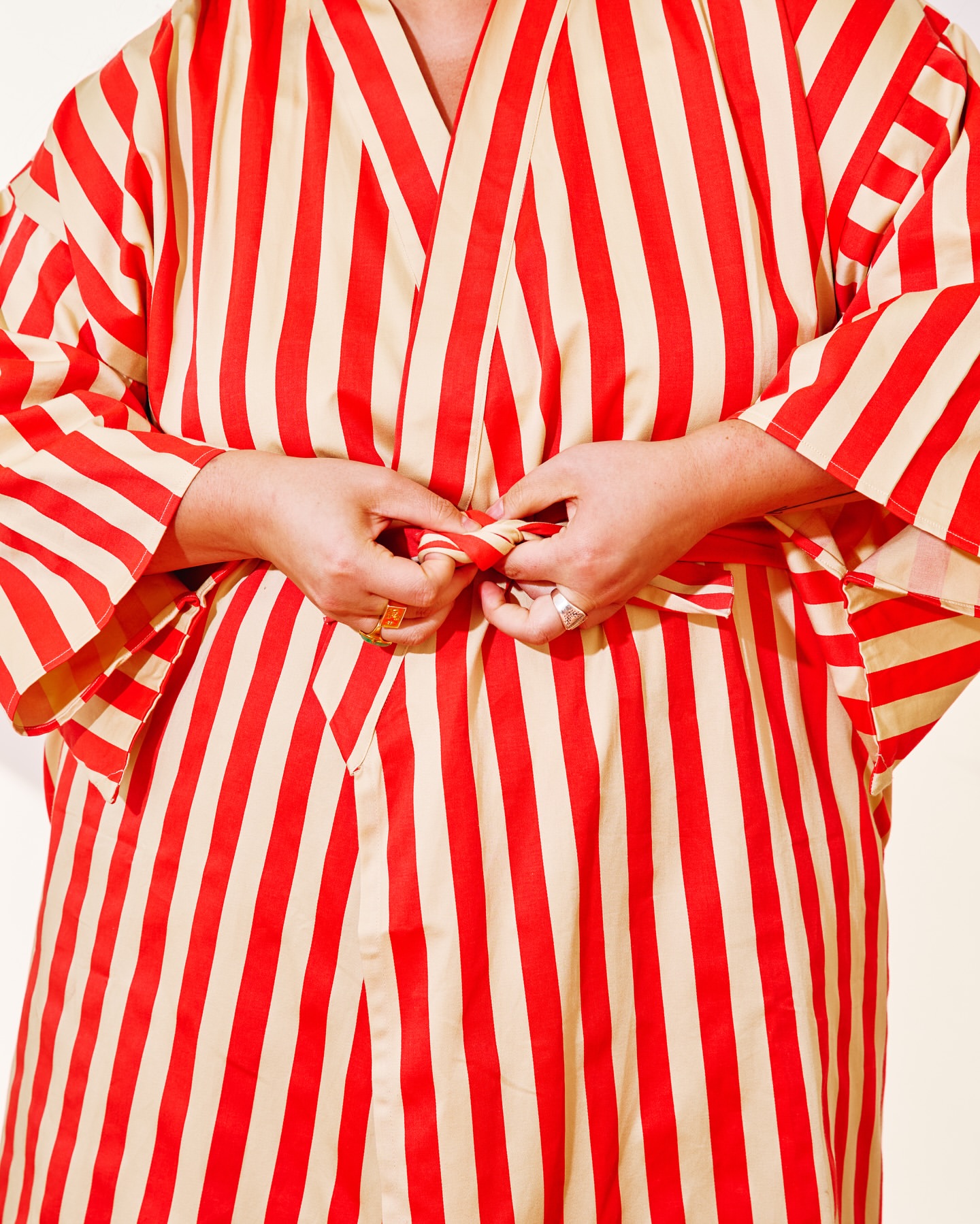 A woman is tying the belt of a red and cream-striped robe with her hands. The close-up image captures only the midsection of the subject.