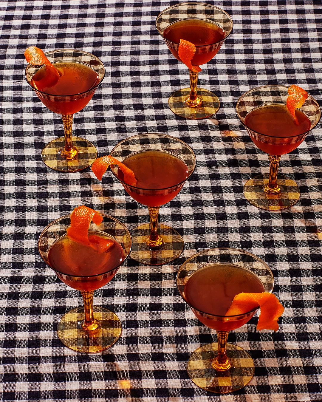 Several stemmed cocktail glasses filled with a dark orange beverage garnished with orange peels on a black and white checkered tablecloth.