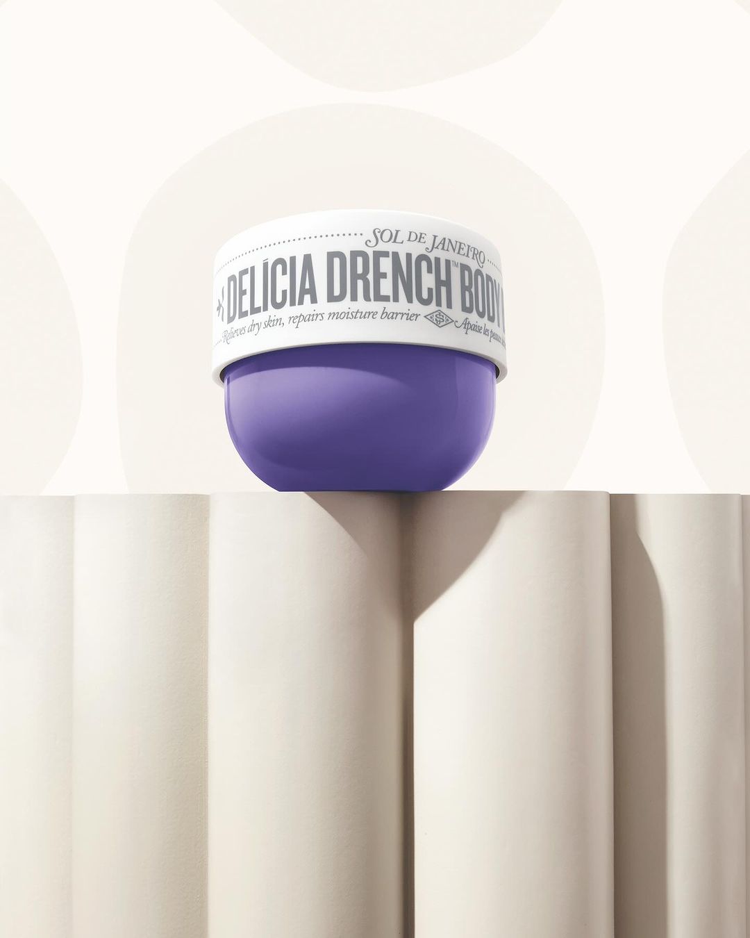 Sol de Janeiro Delicia Drench Body Butter jar in purple and white on a decorative white surface against a cream backdrop.