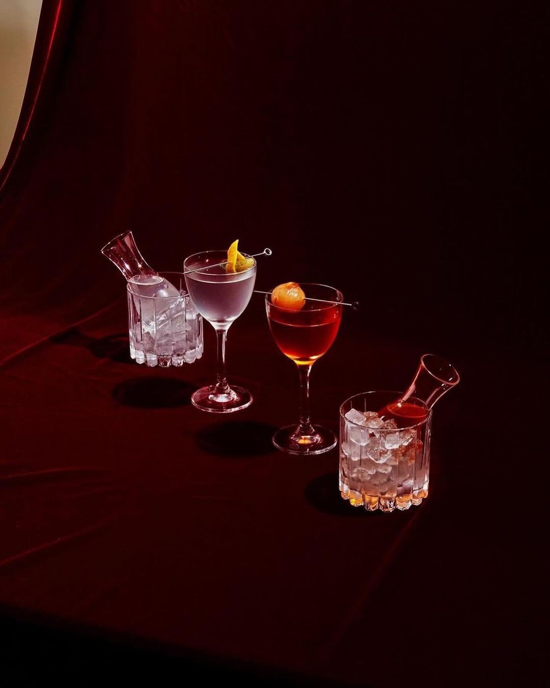 Four cocktails in stemmed glasses and decanters filled with ice sit on a dark red velvety surface against a dark background in a studio setting.