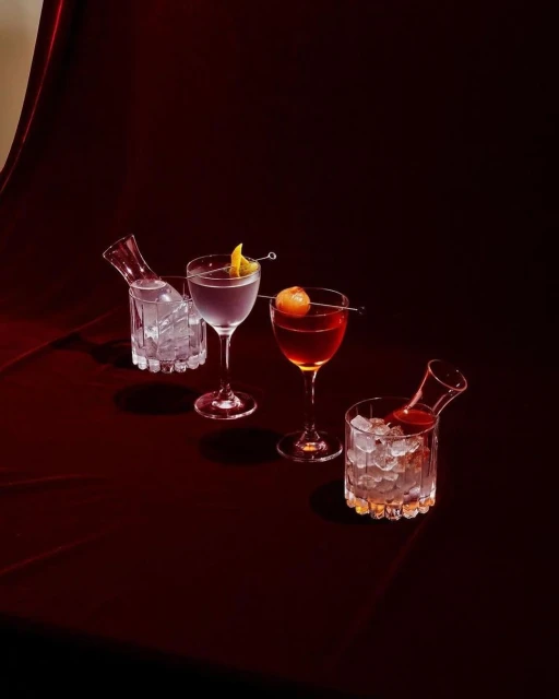 Four cocktails in stemmed glasses and decanters filled with ice sit on a dark red velvety surface against a dark background in a studio setting.