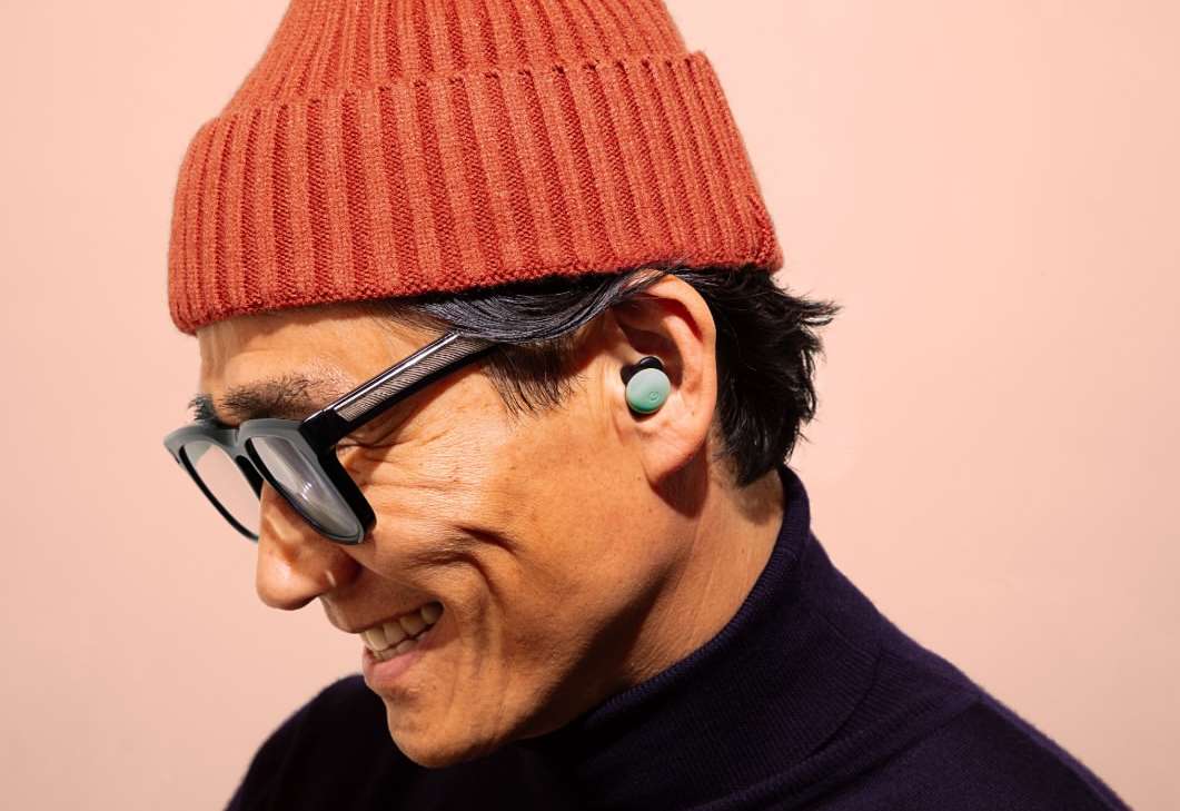 Smiling man wearing glasses, rust-colored knit hat, and wireless earbuds against a light pink background in a close-up shot.
