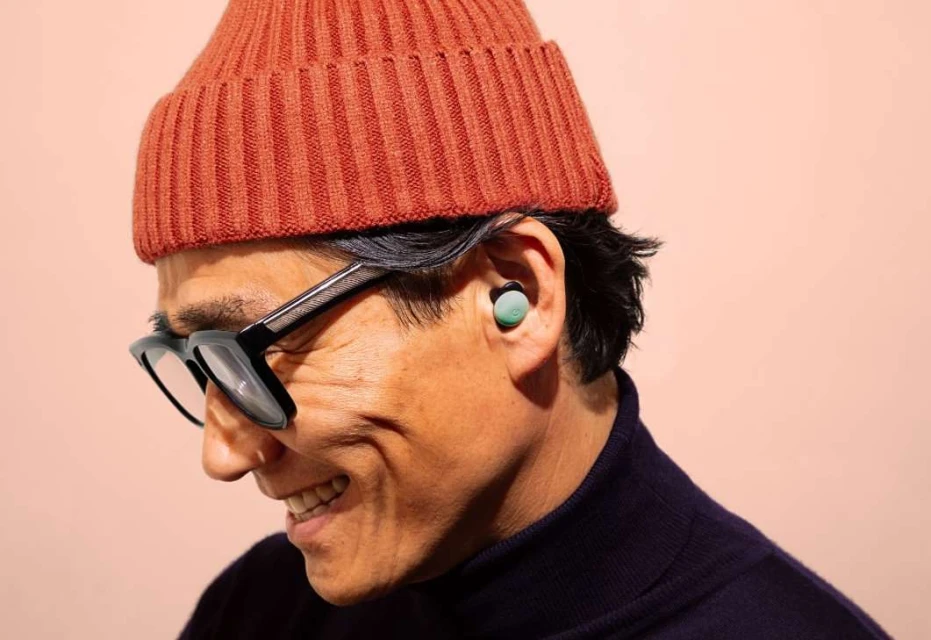 Smiling man wearing glasses, rust-colored knit hat, and wireless earbuds against a light pink background in a close-up shot.