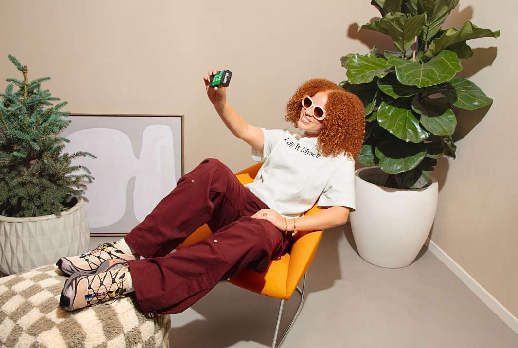 Woman with curly hair wearing sunglasses, sitting in an orange chair and taking a selfie with a green camera indoors with plants.