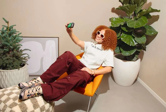 Woman with curly hair wearing sunglasses, sitting in an orange chair and taking a selfie with a green camera indoors with plants.