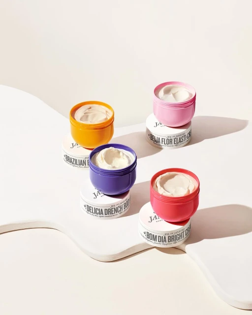 Four colorful Sol de Janeiro body creams are stacked on a white platform. The containers are in pink, orange, purple, and coral.
