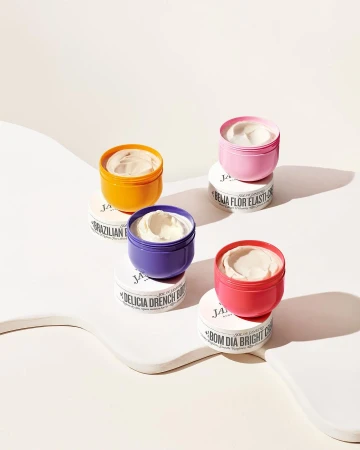 Four colorful Sol de Janeiro body creams are stacked on a white platform. The containers are in pink, orange, purple, and coral.