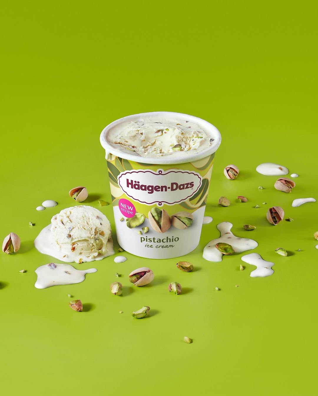 A container of Häagen-Dazs pistachio ice cream sits on a green surface, surrounded by pistachio nuts and melting ice cream drips.