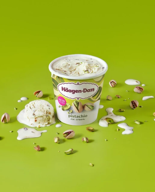 A container of Häagen-Dazs pistachio ice cream sits on a green surface, surrounded by pistachio nuts and melting ice cream drips.