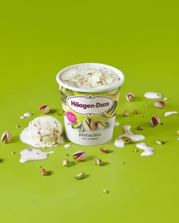 A container of Häagen-Dazs pistachio ice cream sits on a green surface, surrounded by pistachio nuts and melting ice cream drips.