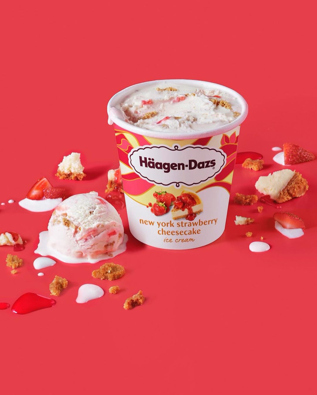 Häagen-Dazs New York Strawberry Cheesecake ice cream tub and a scoop on a red background with strawberry and cake pieces.