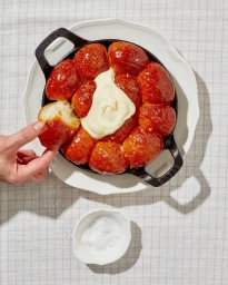 Hand dipping a glazed pull-apart bread roll from a black cast iron skillet topped with cream on a patterned tablecloth