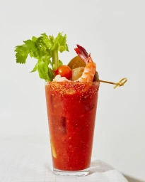 A bloody mary cocktail garnished with celery, cherry tomato, and a skewered shrimp on a light surface.