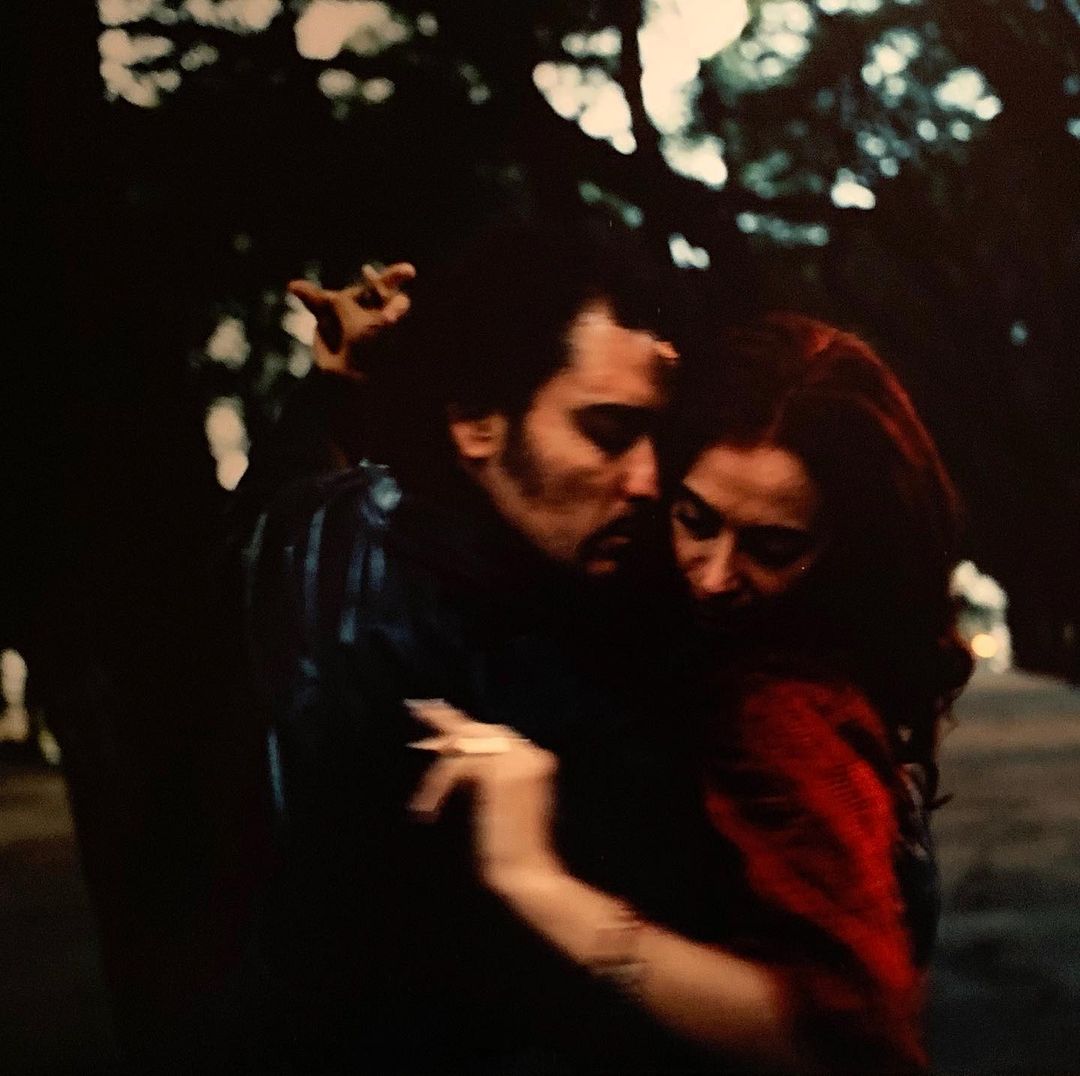 Romantic embrace between a man with a mustache and a woman with red hair in a naturally lit outdoor setting with tree background.
