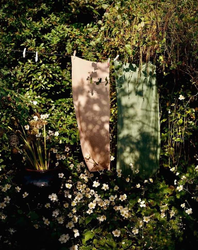 Two skirts hang on a clothesline in a lush garden setting. Soft natural light illuminates the textiles and surrounding foliage.