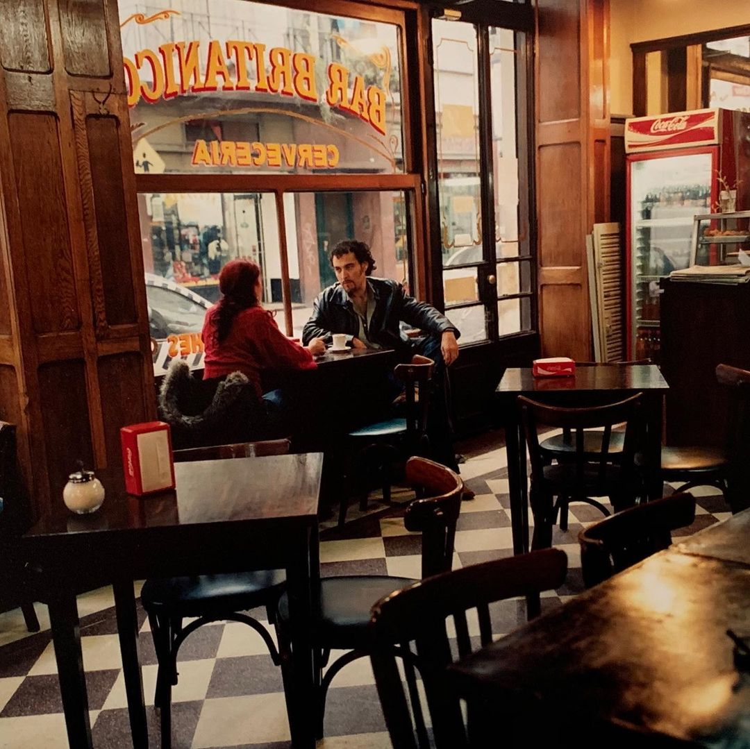 Two people sit at a table inside a café, with dark wooden furniture and a Coca-Cola cooler visible through the window.