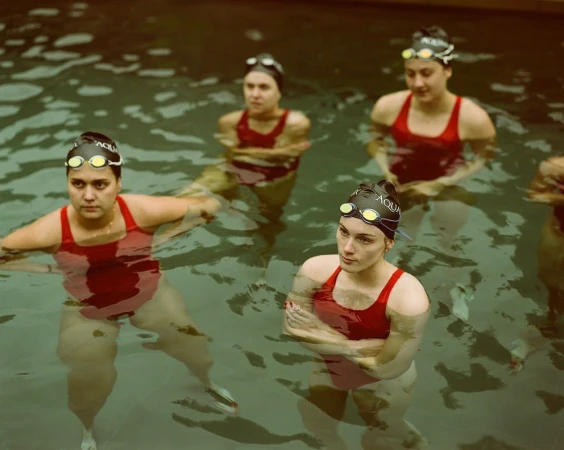 A group of five female athletes in red swimsuits and swim caps are waist-deep in a pool, looking focused and ready for a competition.