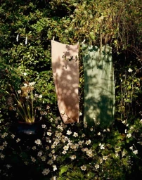 Two hanging fabric panels in beige and green suspended outdoors amidst lush green foliage and flowering plants