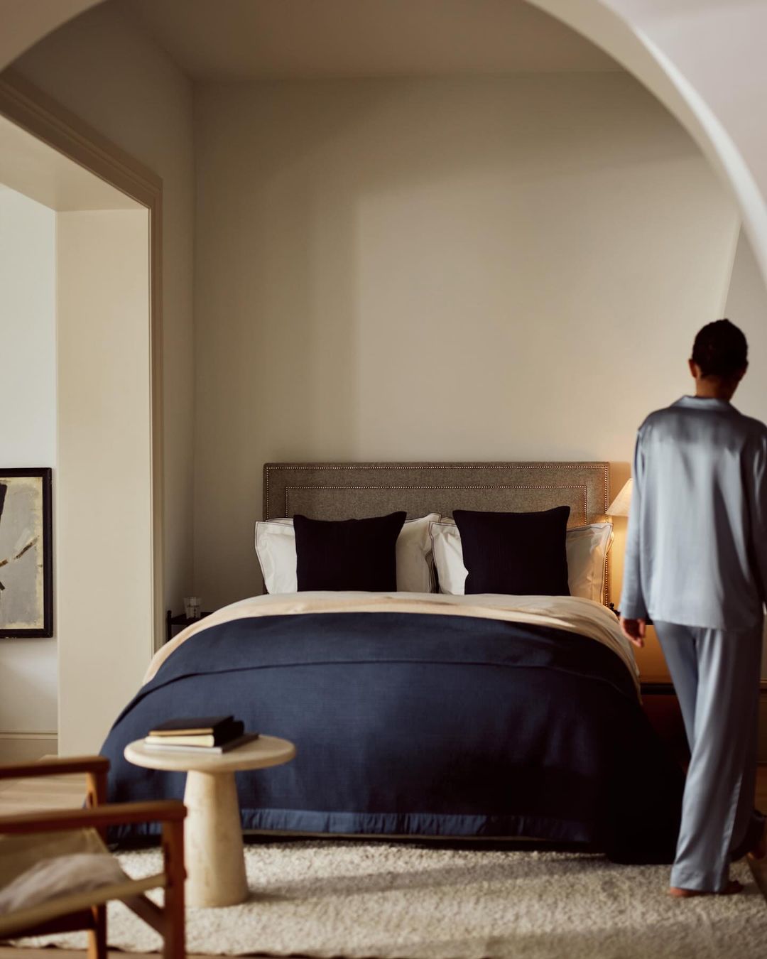 Bedroom interior with a made bed covered with a navy blanket, a side table, and a person entering the room wearing pajamas.