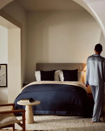 Bedroom interior with a made bed covered with a navy blanket, a side table, and a person entering the room wearing pajamas.