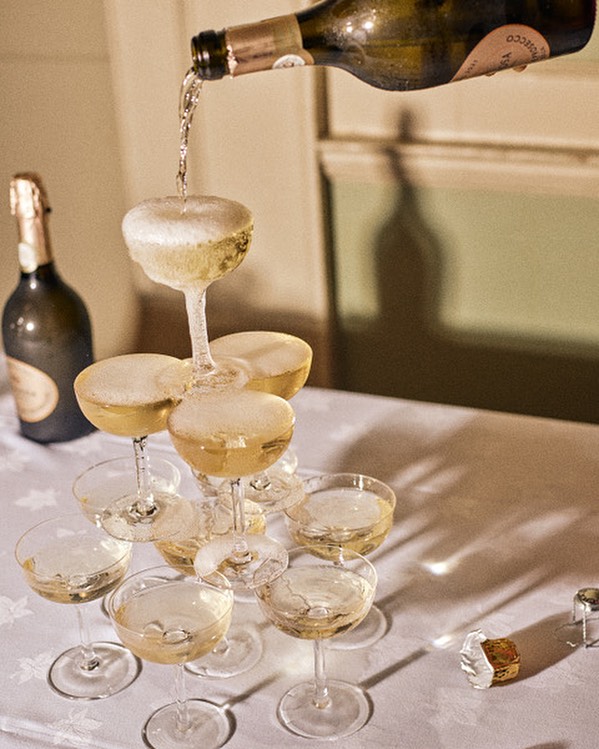 Champagne tower in a stack of coupe glasses on a white tablecloth, liquid being poured from a bottle, indoor celebration setting.