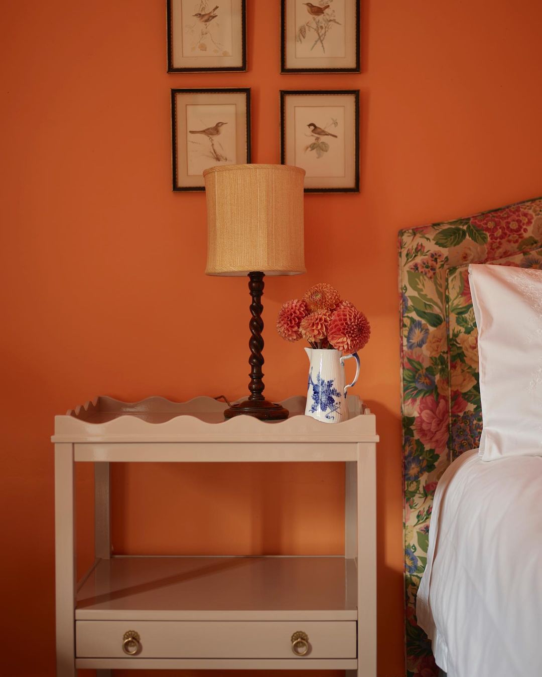 Cozy bedroom interior featuring a nightstand with flowers, a lamp, and artwork on an orange wall, alongside a bed with a floral headboard.