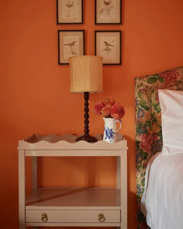 Cozy bedroom interior featuring a nightstand with flowers, a lamp, and artwork on an orange wall, alongside a bed with a floral headboard.