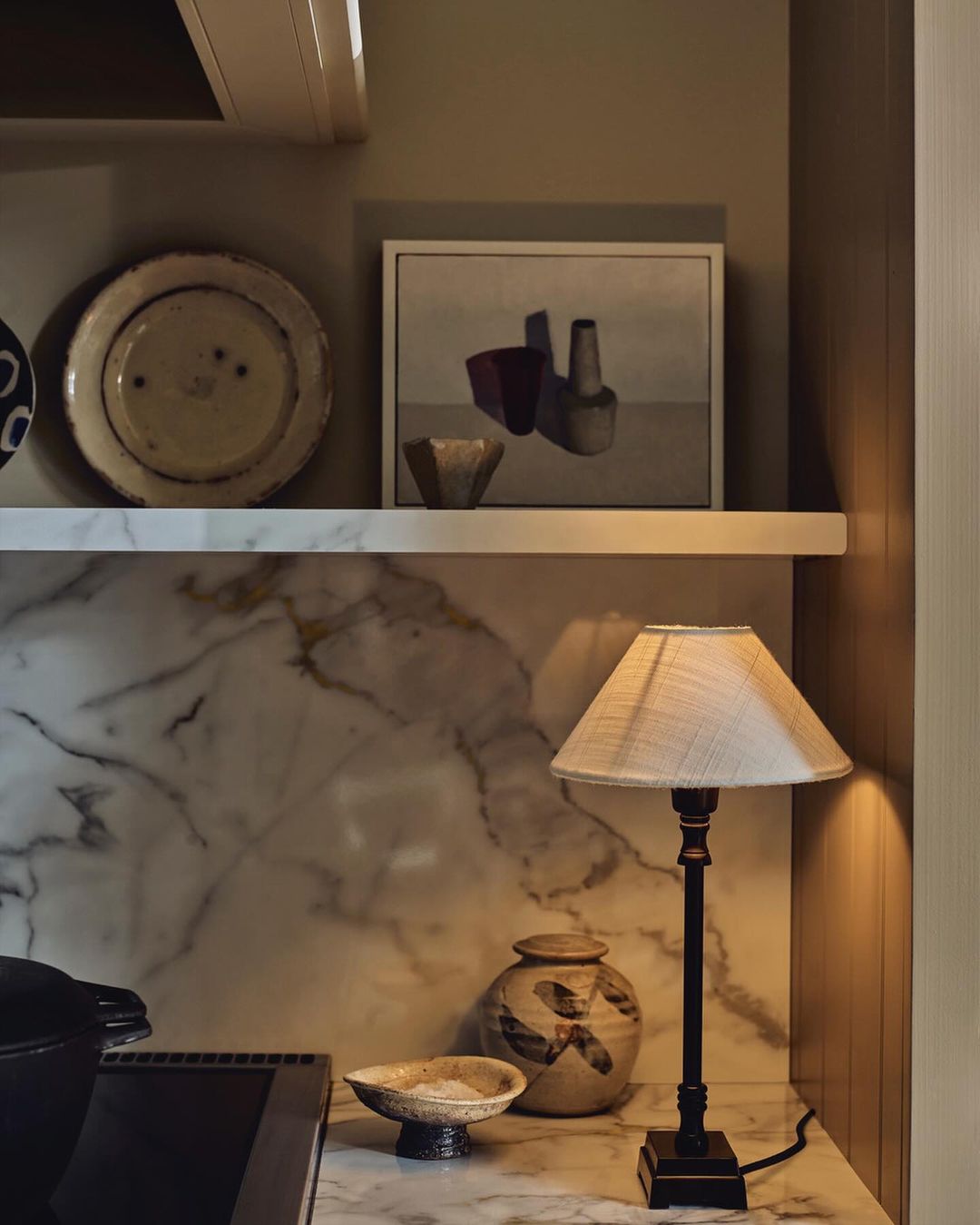 Still life composition featuring a table lamp, ceramic pottery, decorative plates, and framed art, set against a marble backsplash.