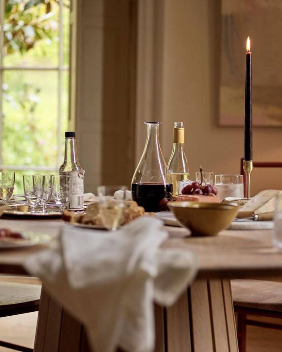 Table setting with wine, grapes, candle, and snacks next to a window providing natural light in a neutral interior space.
