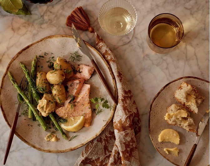 Overhead shot of salmon with potatoes and asparagus plated with bread and wine glasses on a marble surface, showcasing food photography.