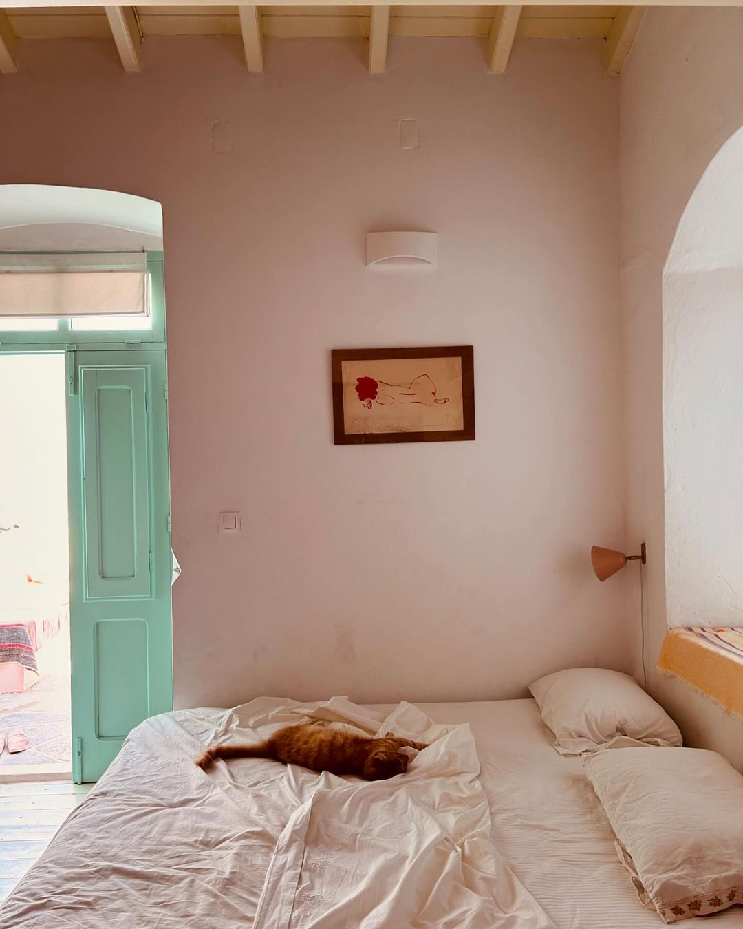 Ginger cat stretches out on white linen bed in a sunlit room with pastel walls, framed art, and open blue door.