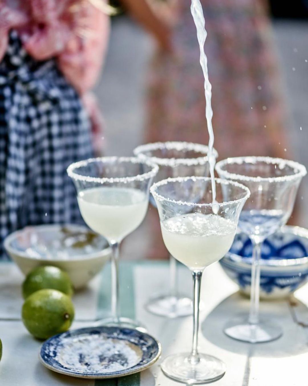 Several margarita cocktails are arranged on a table outside as one is poured. Limes and bowls add decorative elements to the scene.