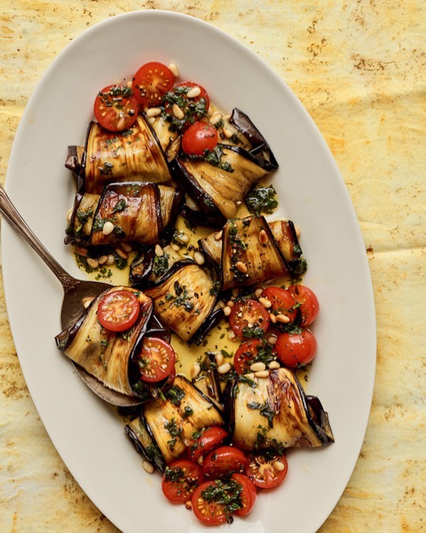 Overhead shot of grilled eggplant rolls with cherry tomatoes, pine nuts, and herbs arranged on a white oval serving platter.