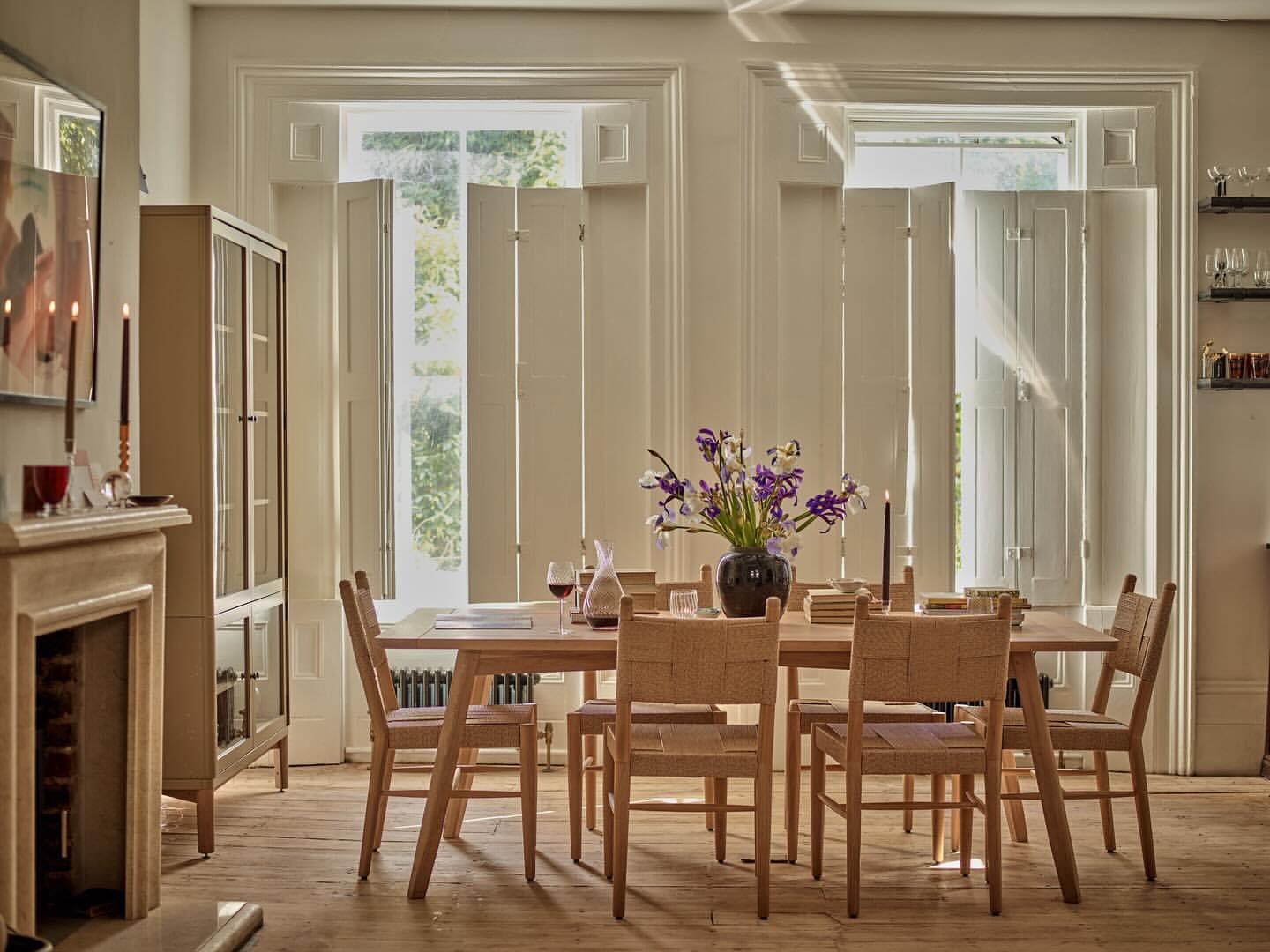 A neutral-toned dining room features a wooden table set with wine glasses and flowers, surrounded by wooden chairs and natural light.