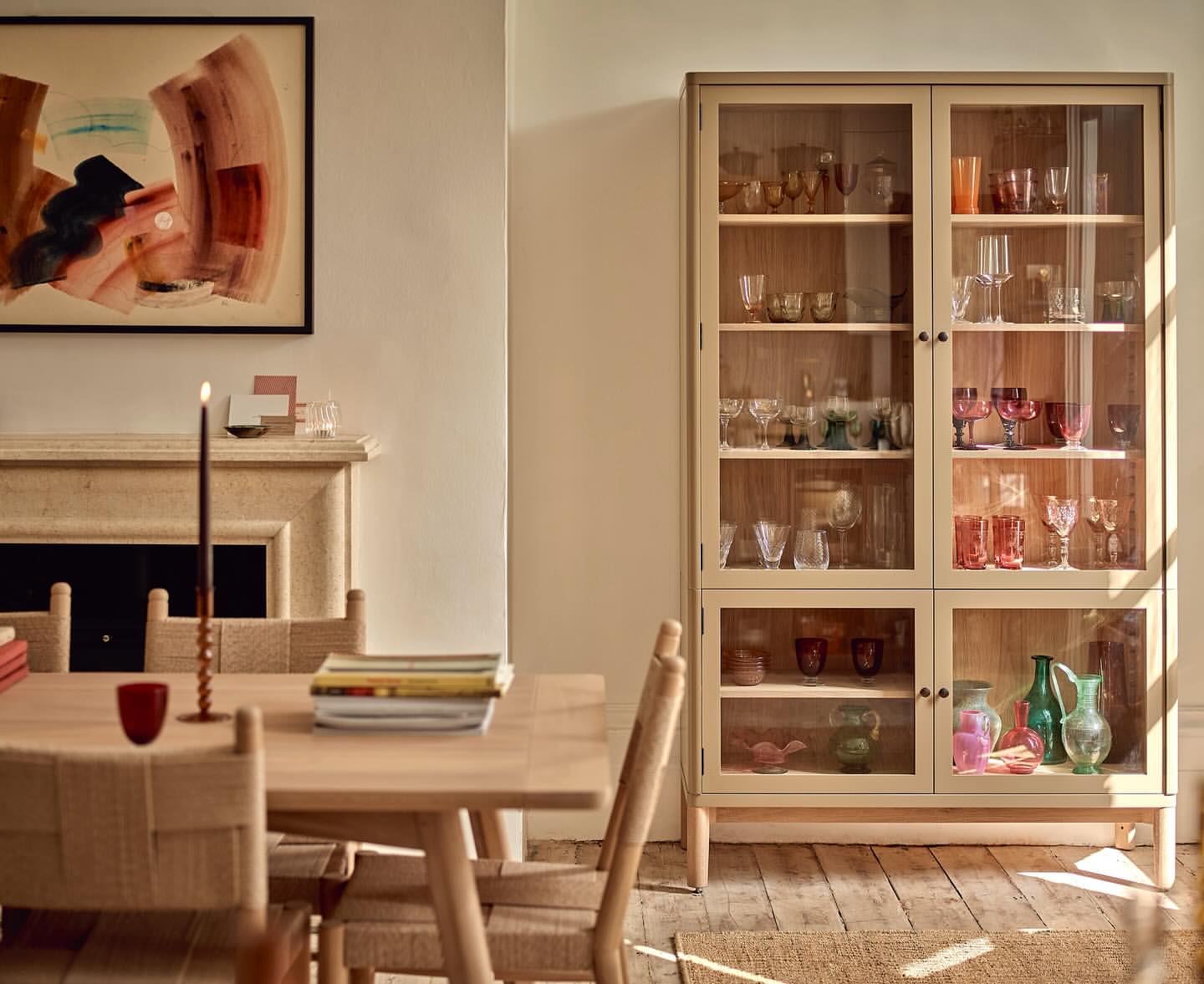 Dining room interior with a glass display cabinet filled with glassware, a wooden table and chairs, and neutral-toned walls.