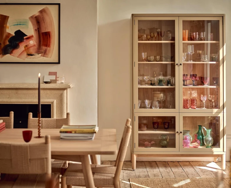 Dining room interior with a glass display cabinet filled with glassware, a wooden table and chairs, and neutral-toned walls.