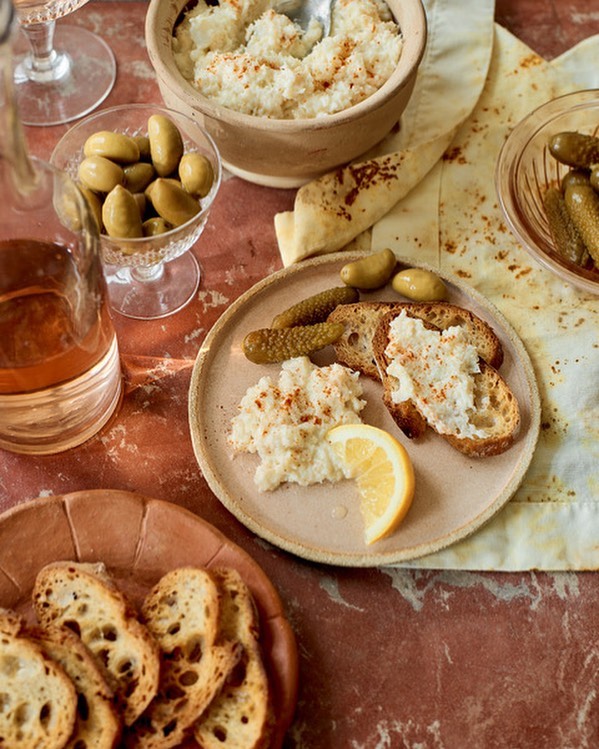 Overhead shot of a food still life with bread, olives, pickles, lemon, and a creamy dip served on plates and bowls on a red marble surface.