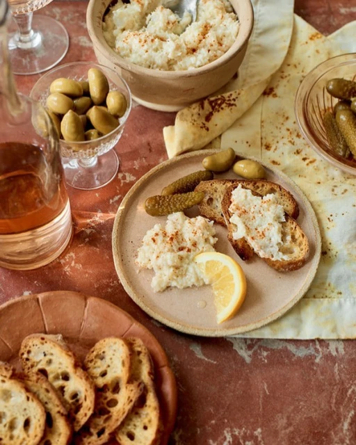 Overhead shot of a food still life with bread, olives, pickles, lemon, and a creamy dip served on plates and bowls on a red marble surface.