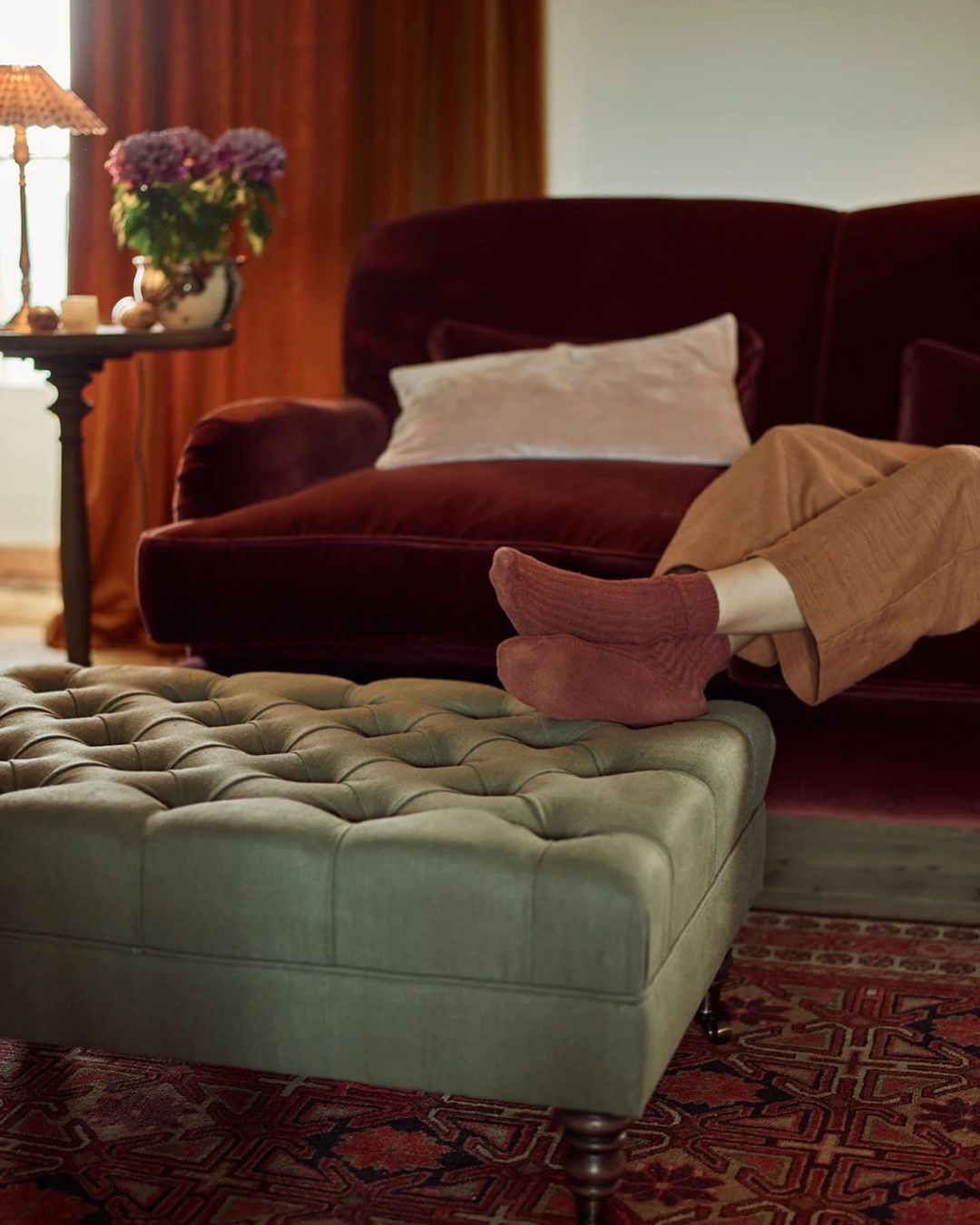 Person relaxing with feet on ottoman, cozy living room interior with velvet sofa, table lamp, and rug, creating a warm, inviting scene.