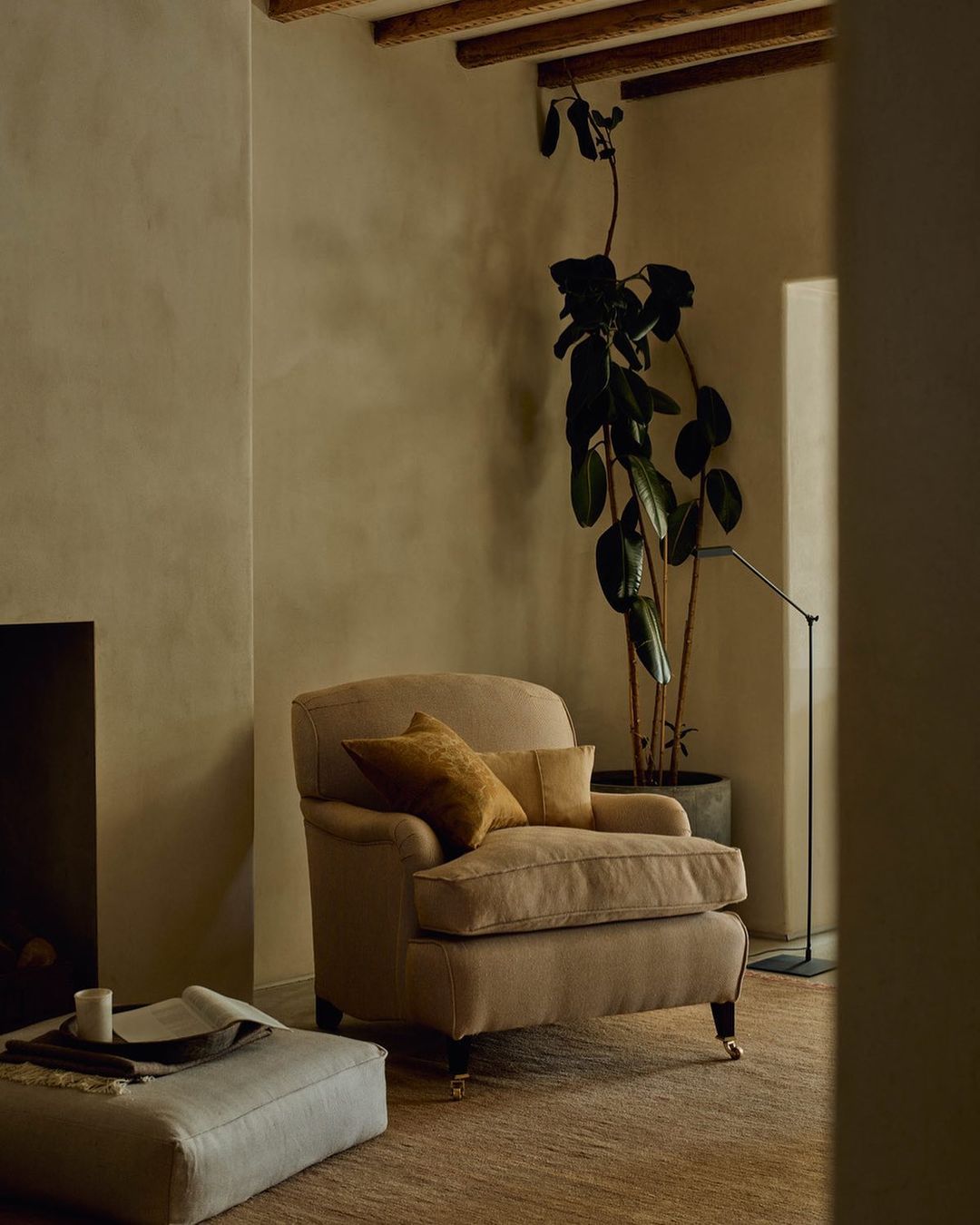 Living room interior with a beige armchair, cushions, floor lamp, houseplant, and an ottoman, creating a cozy and inviting space.