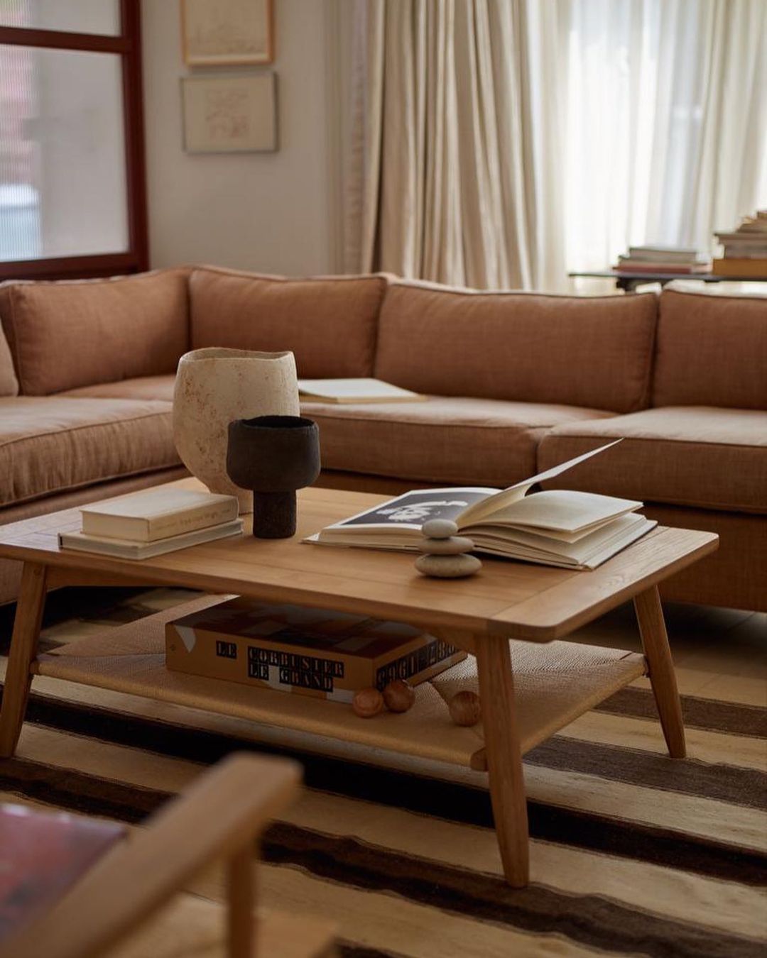 Living room interior featuring a light brown sectional sofa, wooden coffee table with books and decorative objects, and a striped rug.