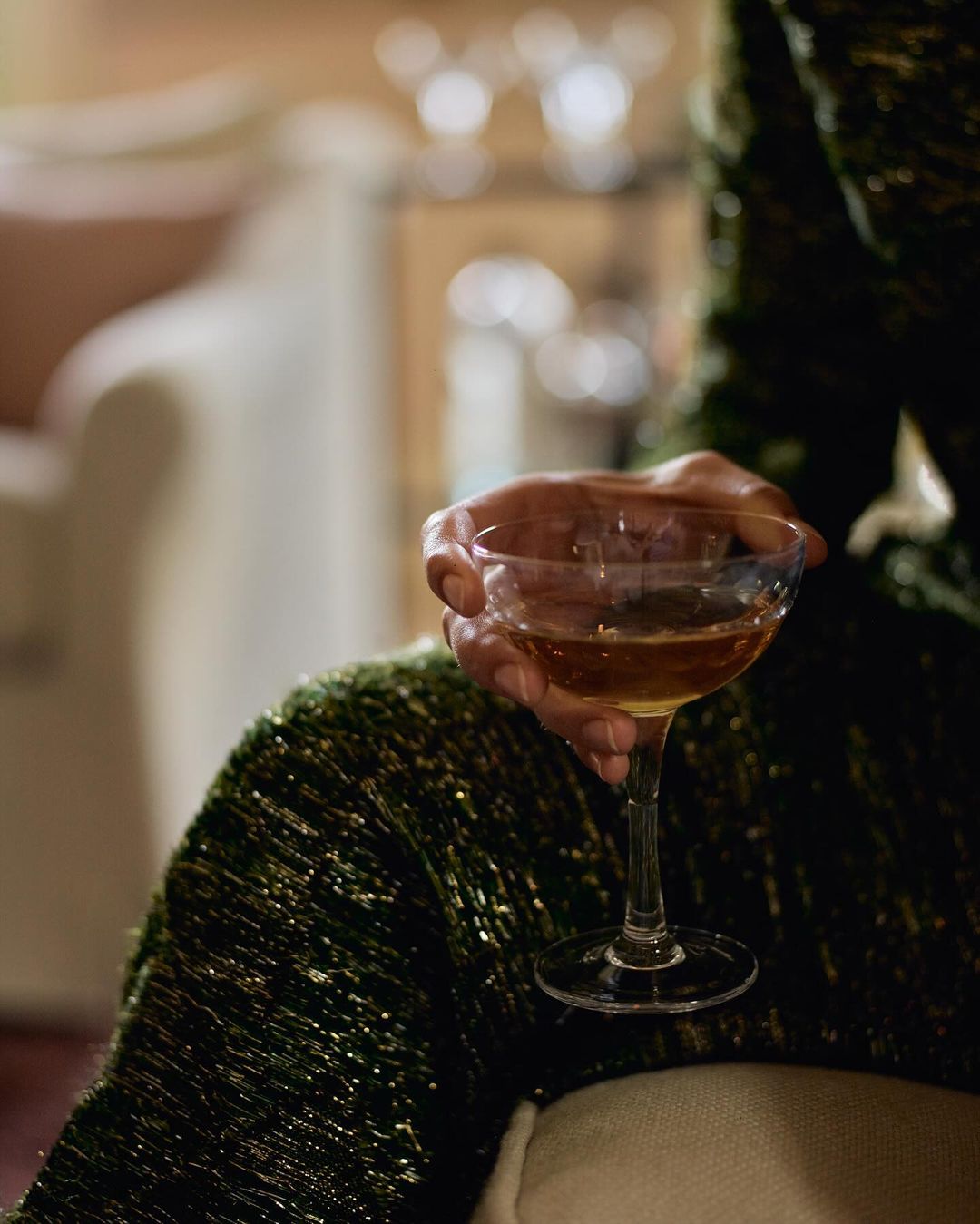 A person holds a stemmed cocktail glass filled with a brown liquid, wearing a sparkly green jacket in an indoor setting with soft lighting.