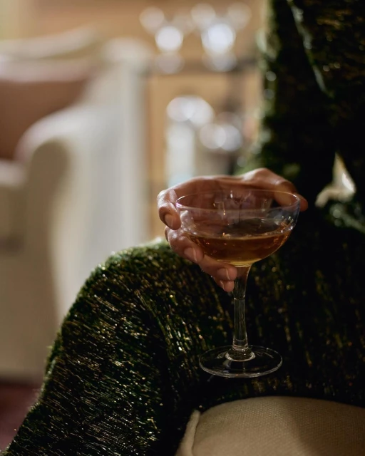 A person holds a stemmed cocktail glass filled with a brown liquid, wearing a sparkly green jacket in an indoor setting with soft lighting.