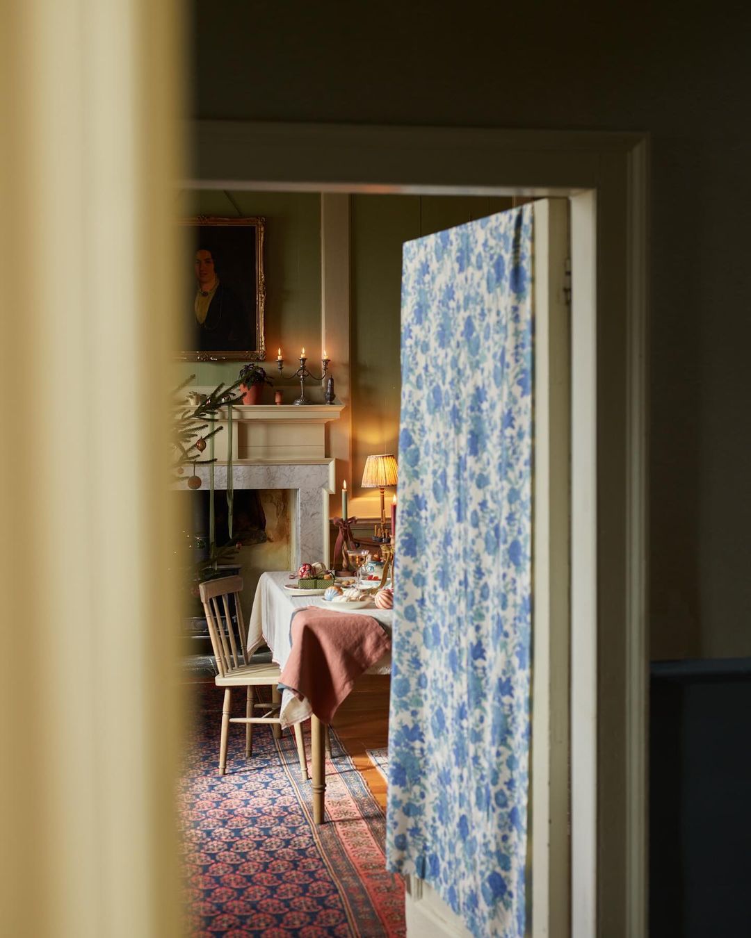 Warm-toned dining room featuring a table set with dishes, candles, and a painting above the fireplace, viewed through a doorway.