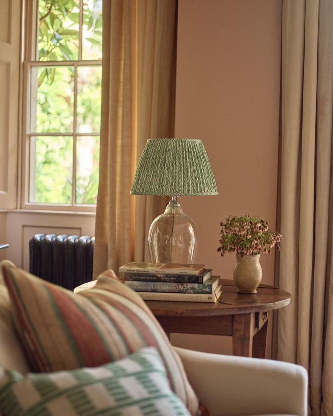 Interior with clear glass lamp and green pleated shade on stack of books on wooden table next to sofa, drapes, and window.