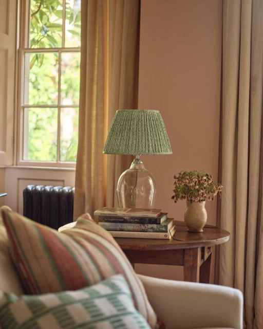 Interior with clear glass lamp and green pleated shade on stack of books on wooden table next to sofa, drapes, and window.