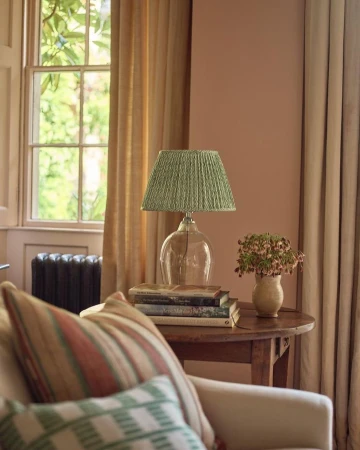 Interior with clear glass lamp and green pleated shade on stack of books on wooden table next to sofa, drapes, and window.