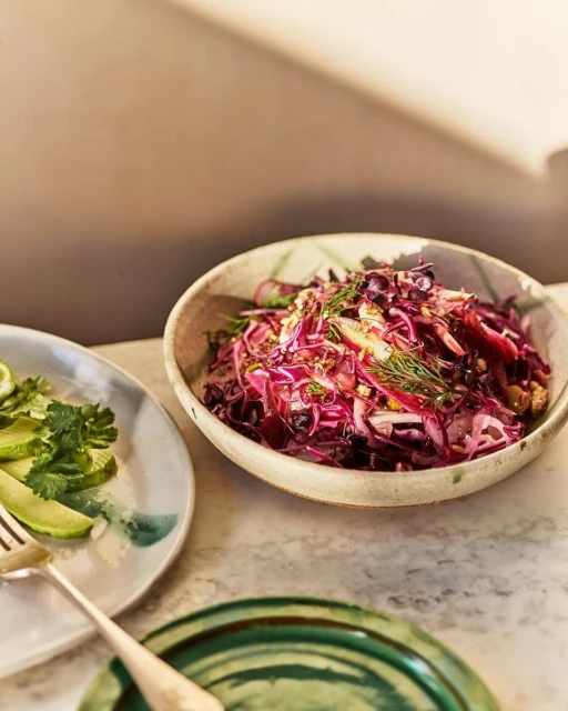 Overhead shot of red cabbage salad in a ceramic bowl, with plates of lime slices and herbs on a marble surface in natural light.