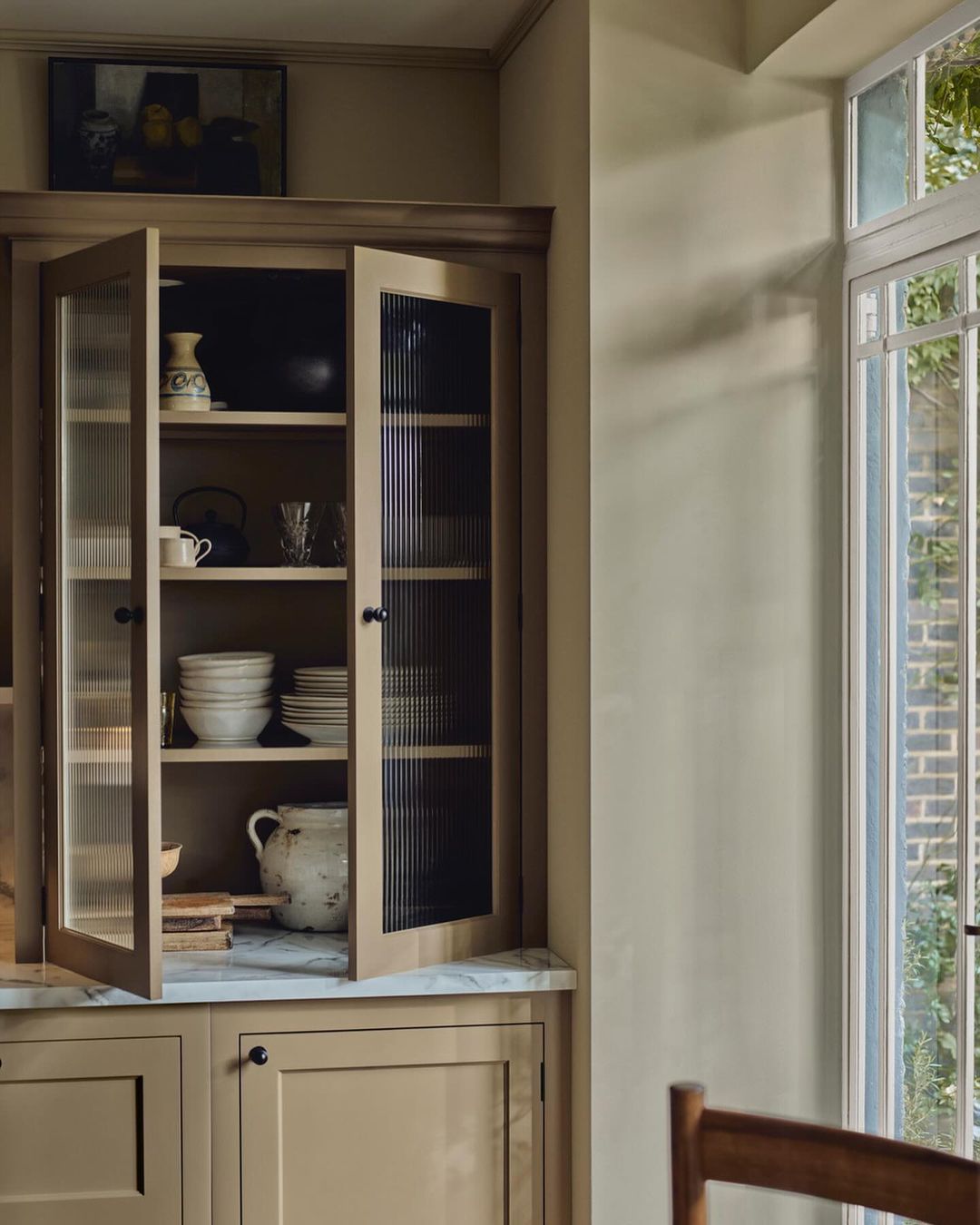 Interior shot of a kitchen hutch with glass doors revealing dishware, set against a neutral-toned wall near a bright window.