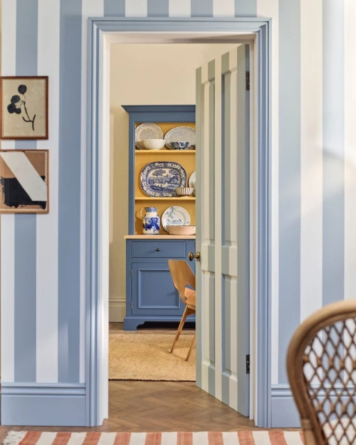 Interior view of a dining room with light blue striped walls, a vintage cabinet with dishware, and an open doorway.